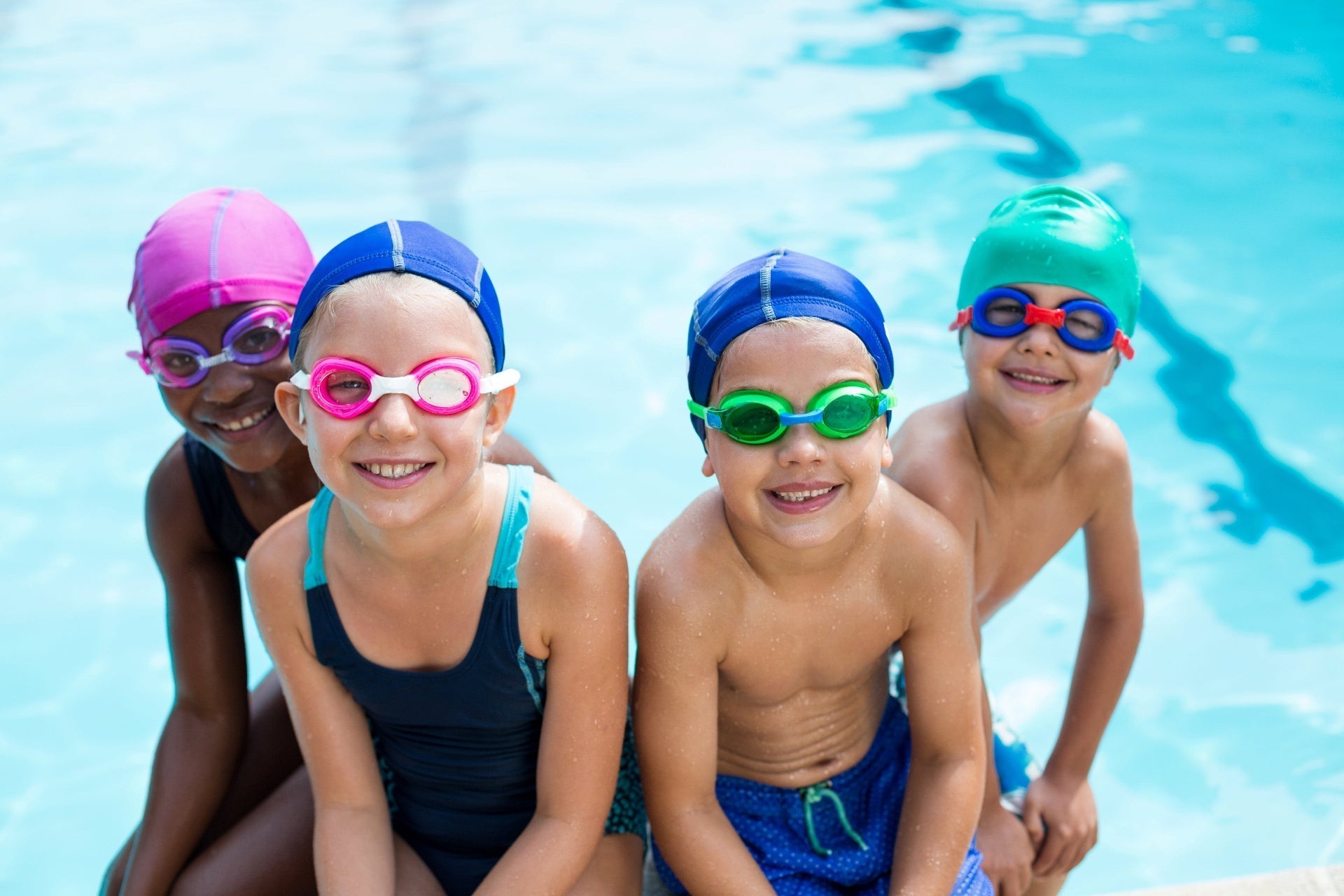 Four smiling children sitting by a swimming pool wearing goggles to protecttheir eyes, swim caps to protect their hair, and barrier lotion to protect their skin