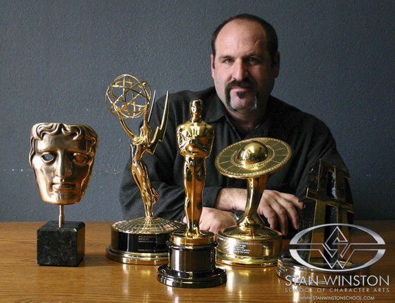 Howard Berger posing with various awards including Oscars and Emmys on a wooden surface.