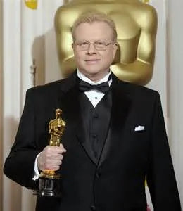Greg Cannom in a black tuxedo holding an Oscar statuette, standing in front of a large gold Oscar sculpture.